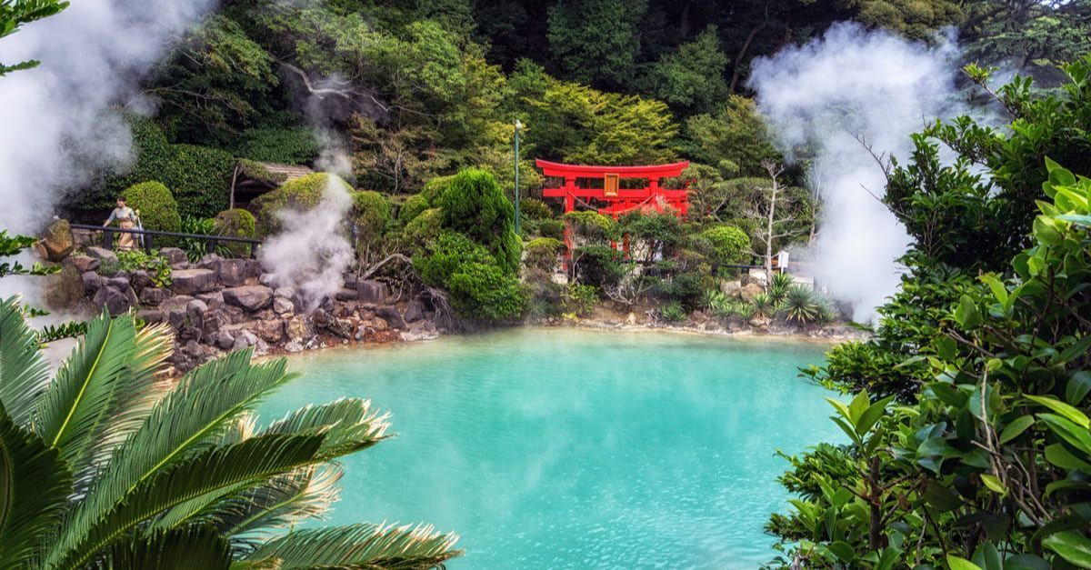 Vapore si innalza da una sorgente termale turchese in una foresta lussureggiante con un cancello torii giapponese rosso sullo sfondo.