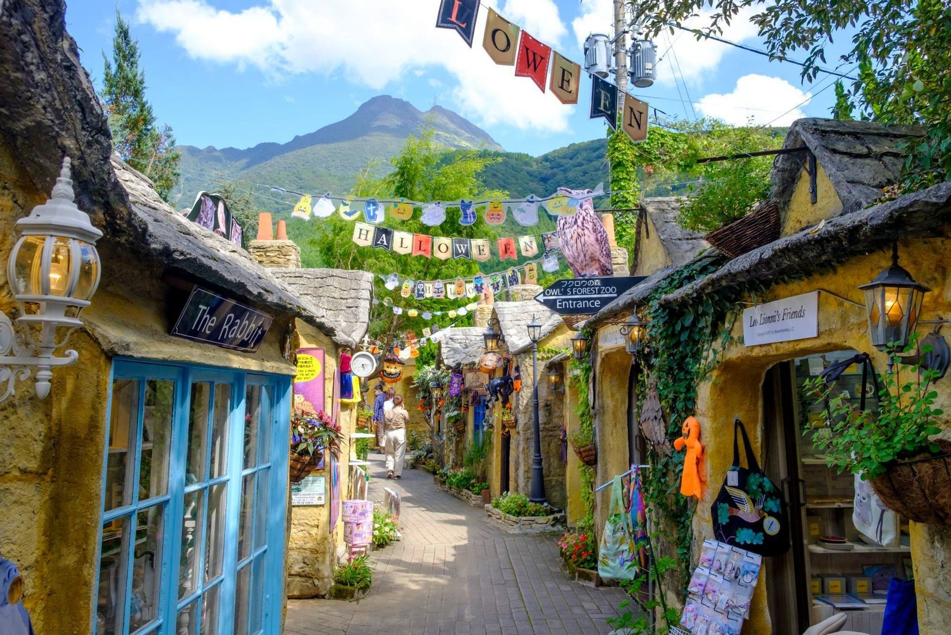 Una stretta strada acciottolata in un villaggio fantasy con negozi decorati per Halloween e una montagna visibile sullo sfondo.