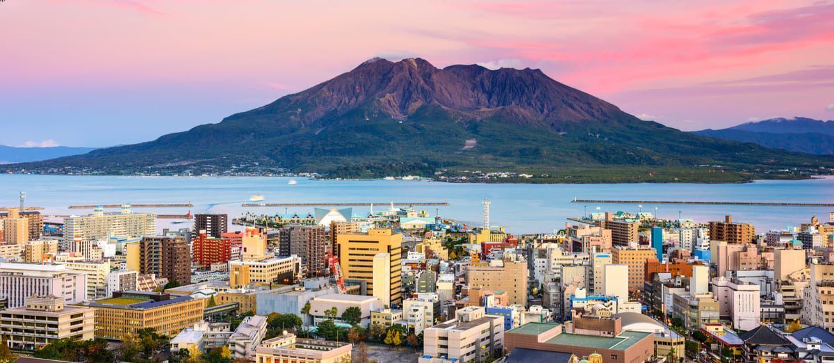 Una vista panoramica di una città costiera con un grande vulcano di fronte alla baia sotto un cielo rosa al tramonto.