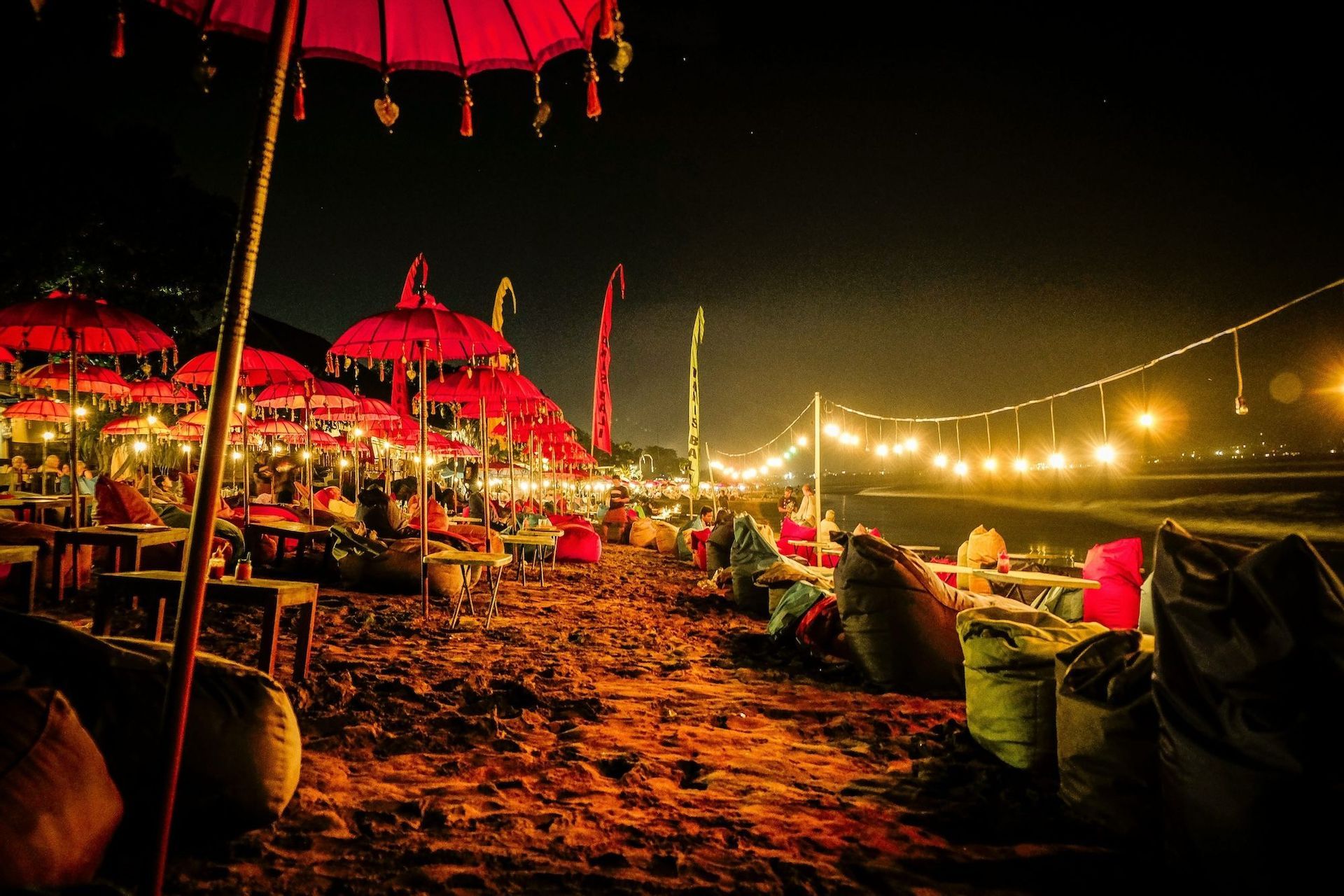 Fila di ombrelloni rossi e pouf su una spiaggia sabbiosa di notte, illuminati da calde lucine.