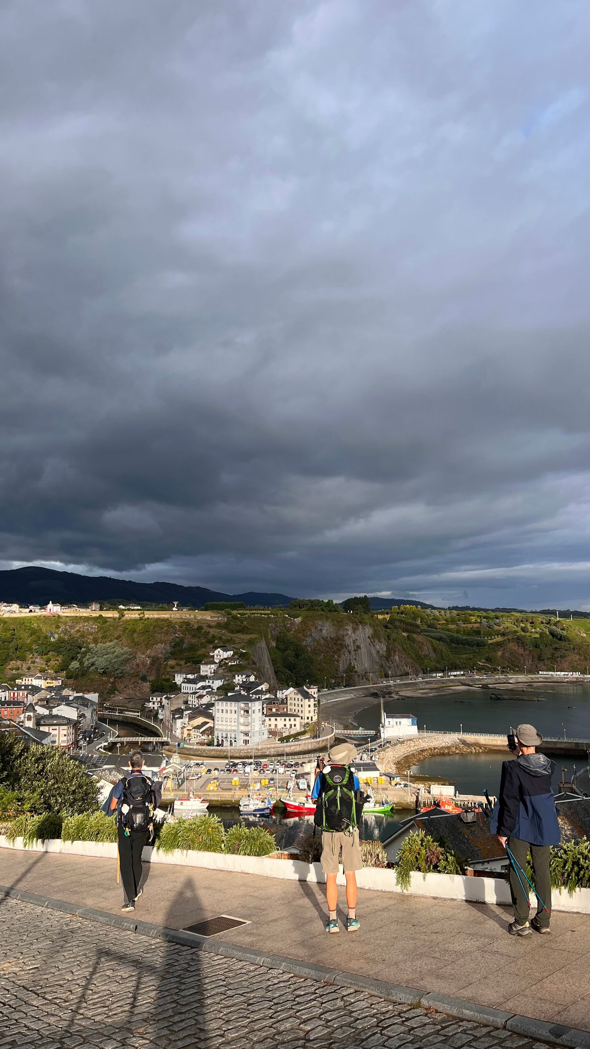 Un gruppo WeRoad di tre escursionisti con zaini che domina una cittadina portuale costiera da un punto panoramico sotto un cielo nuvoloso e drammatico.