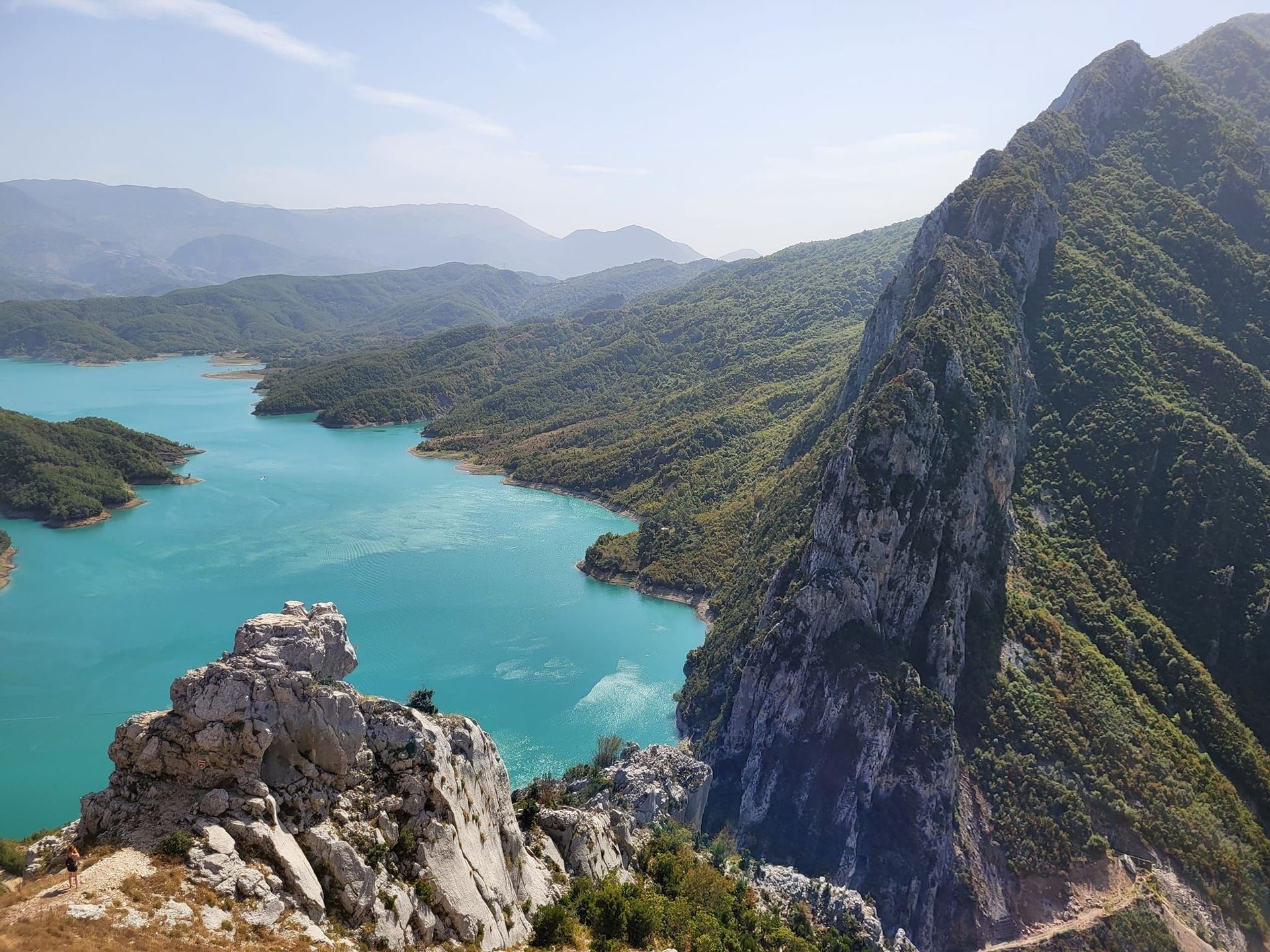 Una vista dall'alto da un crinale montuoso roccioso che si affaccia su un sinuoso lago turchese circondato da verdi colline boscose.