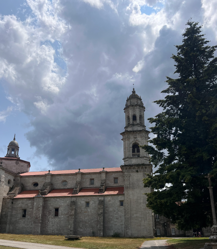 Una chiesa in pietra con campanile e tetto in tegole rosse si erge accanto a un alto albero sempreverde sotto un cielo drammatico e nuvoloso.