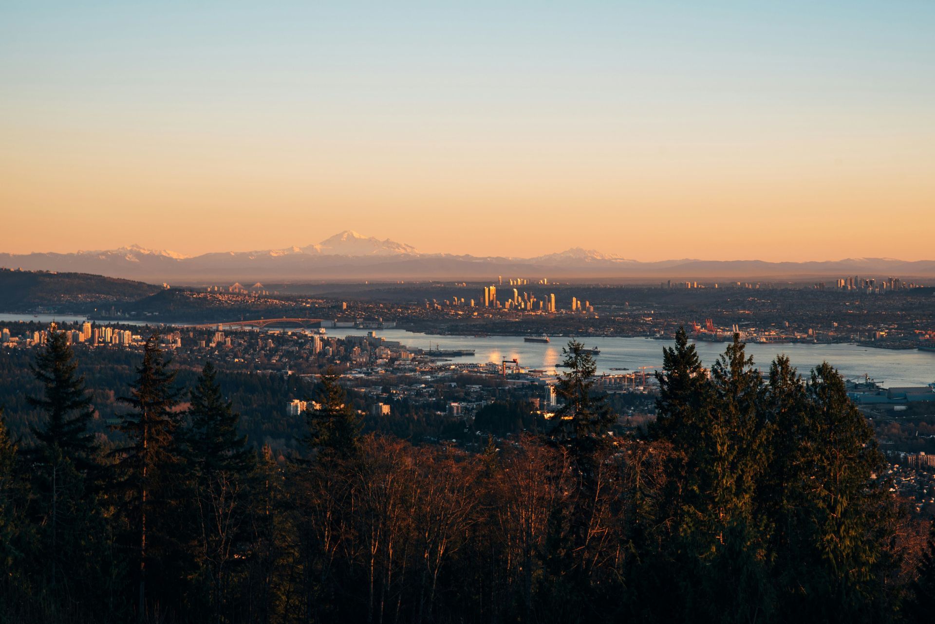 Vue plongeante sur une silhouette urbaine au bord de l'eau au lever du soleil, avec des montagnes enneigées au loin et la cime des arbres en contrebas.