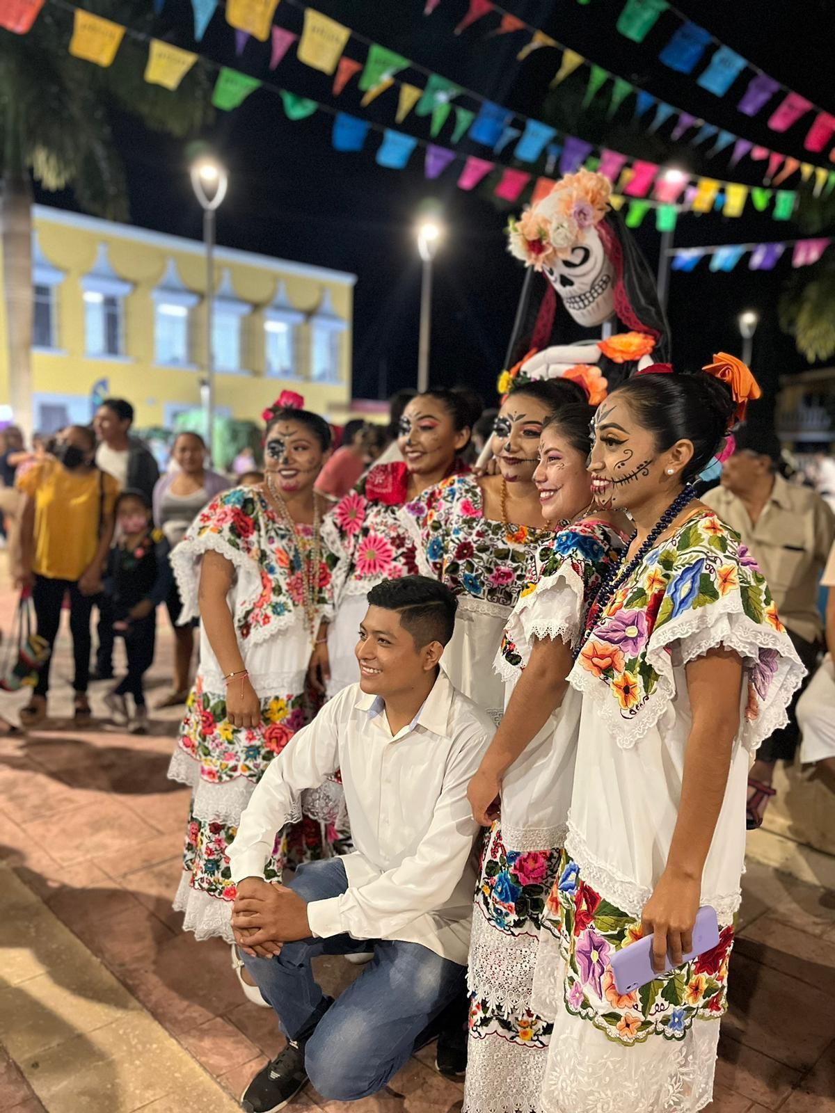 Un gruppo di persone in abiti tradizionali e trucco da Catrina posa per una foto a un festival notturno sotto bandiere colorate.