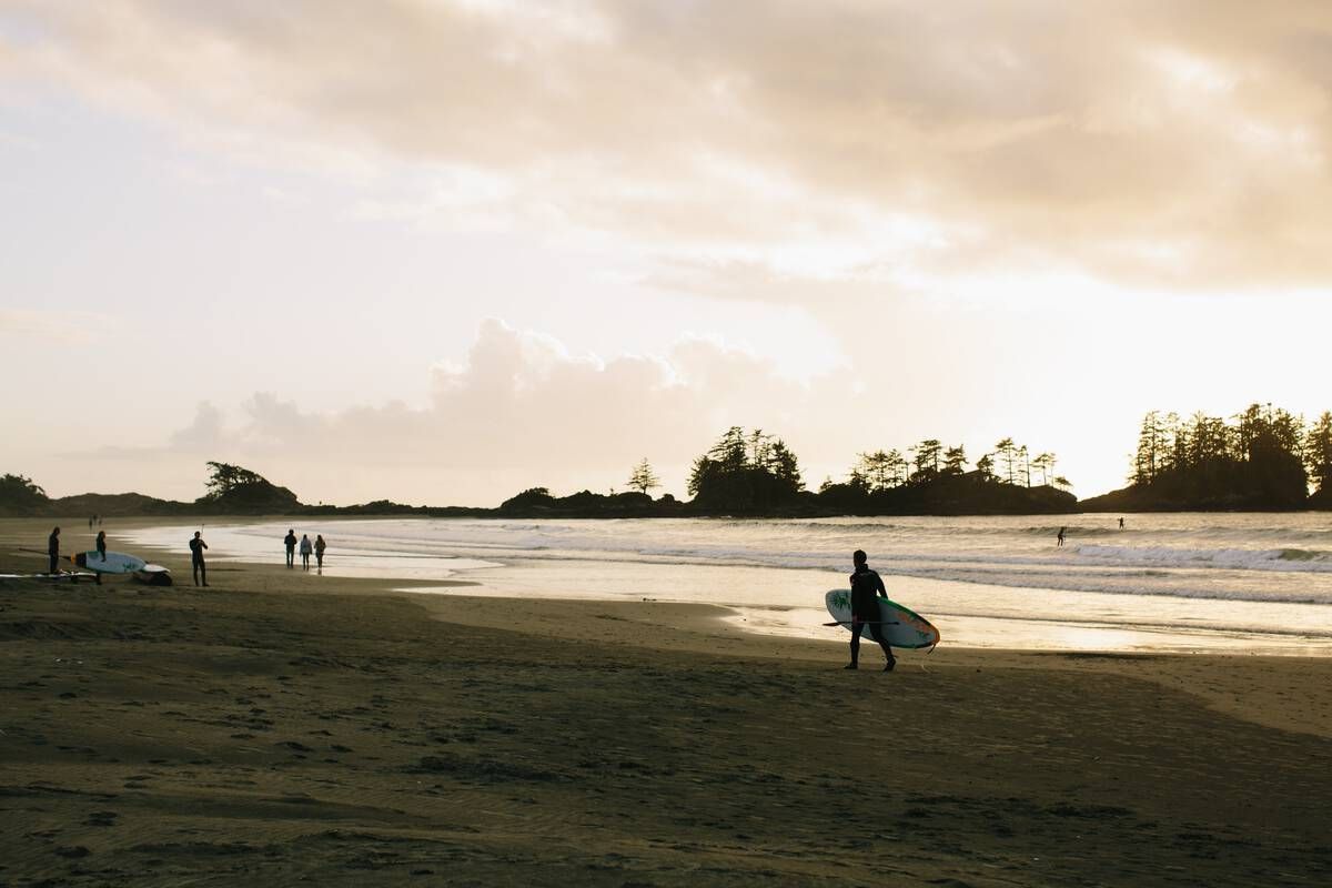 Une personne en combinaison de plongée porte une planche de surf le long d'une plage de sable au coucher du soleil, avec d'autres surfeurs en arrière-plan.