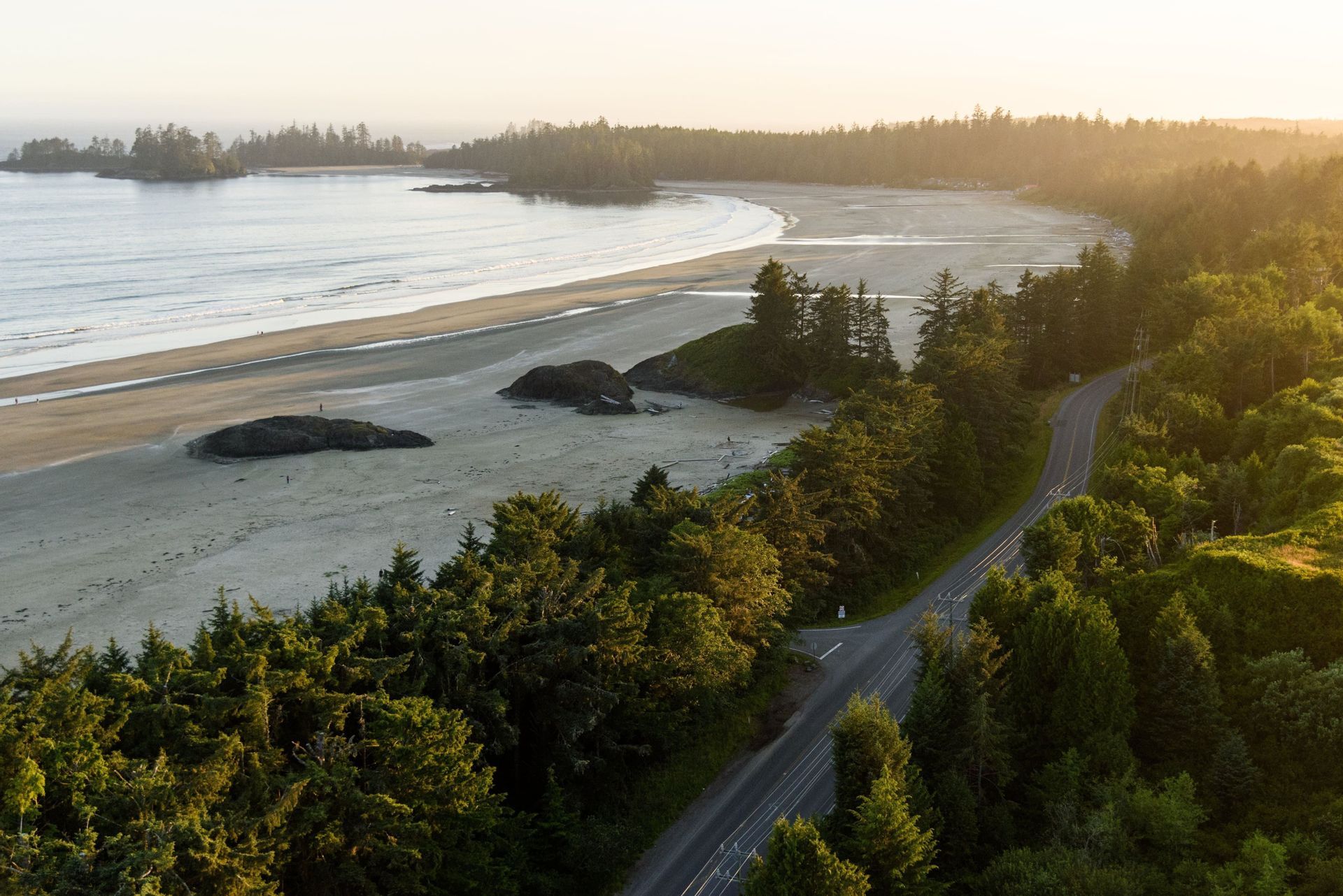 Vue aérienne d'une route sinueuse traversant une forêt luxuriante et verdoyante le long d'une large plage de sable au coucher du soleil.