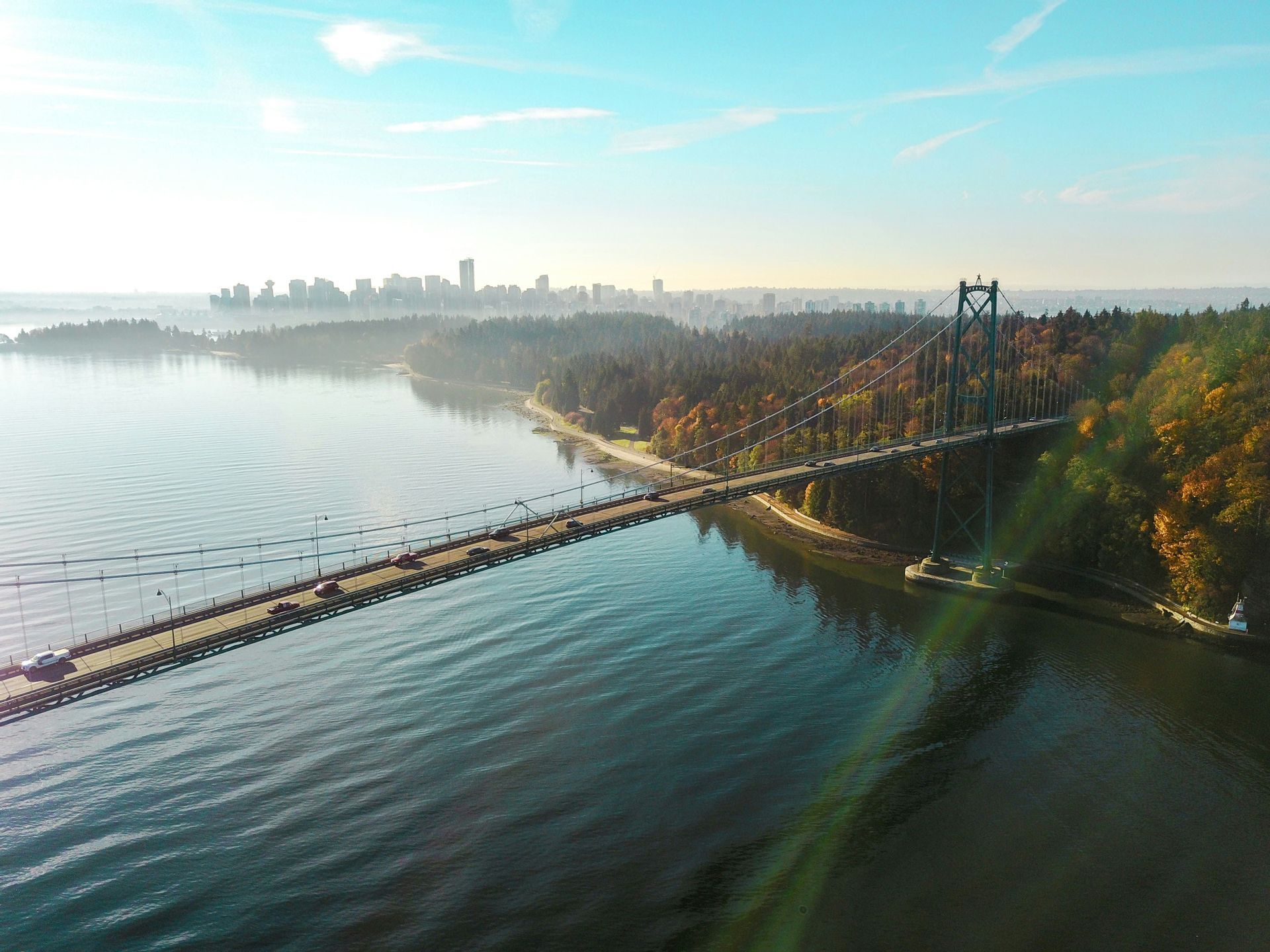 Vue aérienne d'un pont suspendu avec des voitures roulant sur l'eau, reliant un littoral boisé à l'horizon d'une ville lointaine.