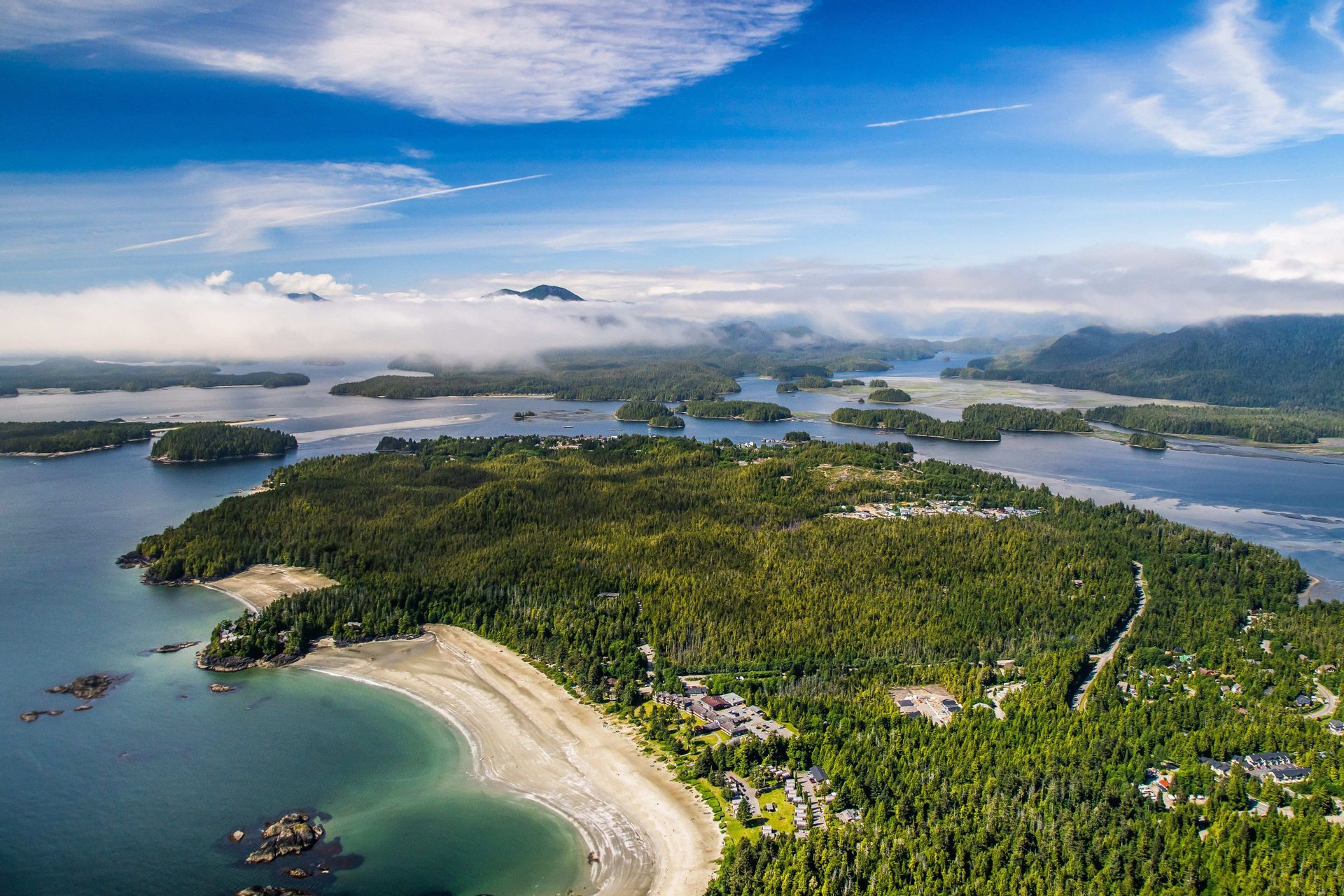 Vue aérienne d'un archipel boisé avec une plage de sable incurvée, entouré d'eau et de petites îles sous un ciel partiellement nuageux.