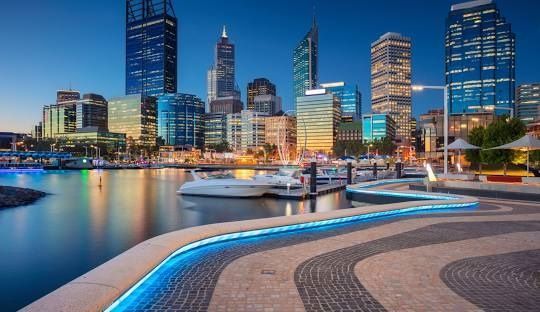 Un horizonte urbano moderno con rascacielos iluminados al anochecer, reflejándose en un puerto deportivo con barcos amarrados y un paseo marítimo curvo e iluminado.