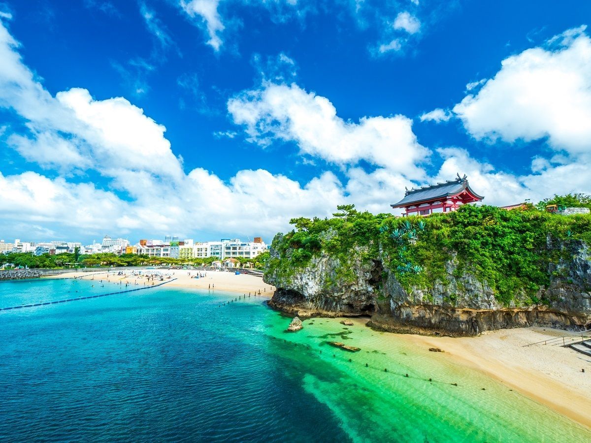 Un santuario giapponese con un tetto rosso si erge su una scogliera rocciosa e verde sopra una spiaggia sabbiosa con acqua turchese sotto un cielo azzurro.