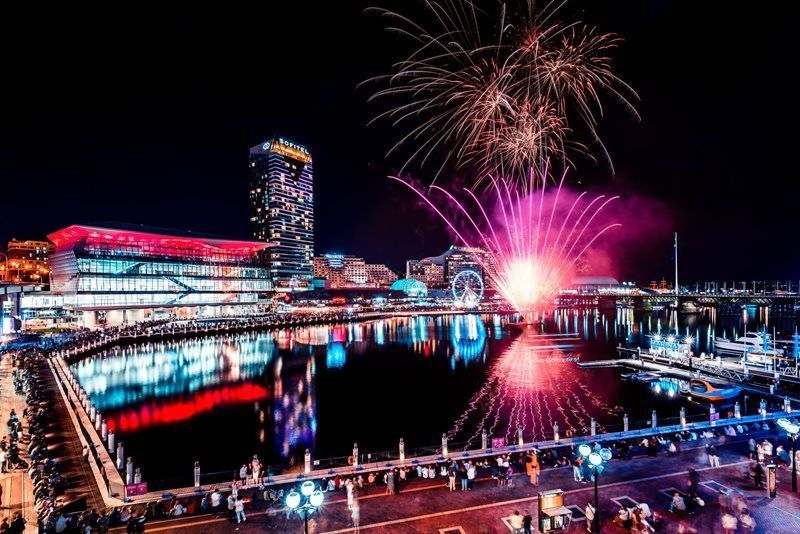 Fuegos artificiales rosas estallan sobre un moderno puerto de la ciudad por la noche, con multitudes observando desde el muelle y luces reflejándose en el agua.
