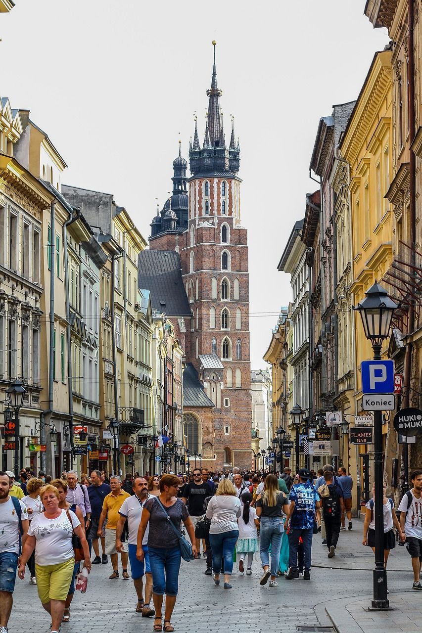 Una multitud de personas camina por una estrecha calle histórica, flanqueada por edificios antiguos, con una gran torre de iglesia de ladrillo al fondo.