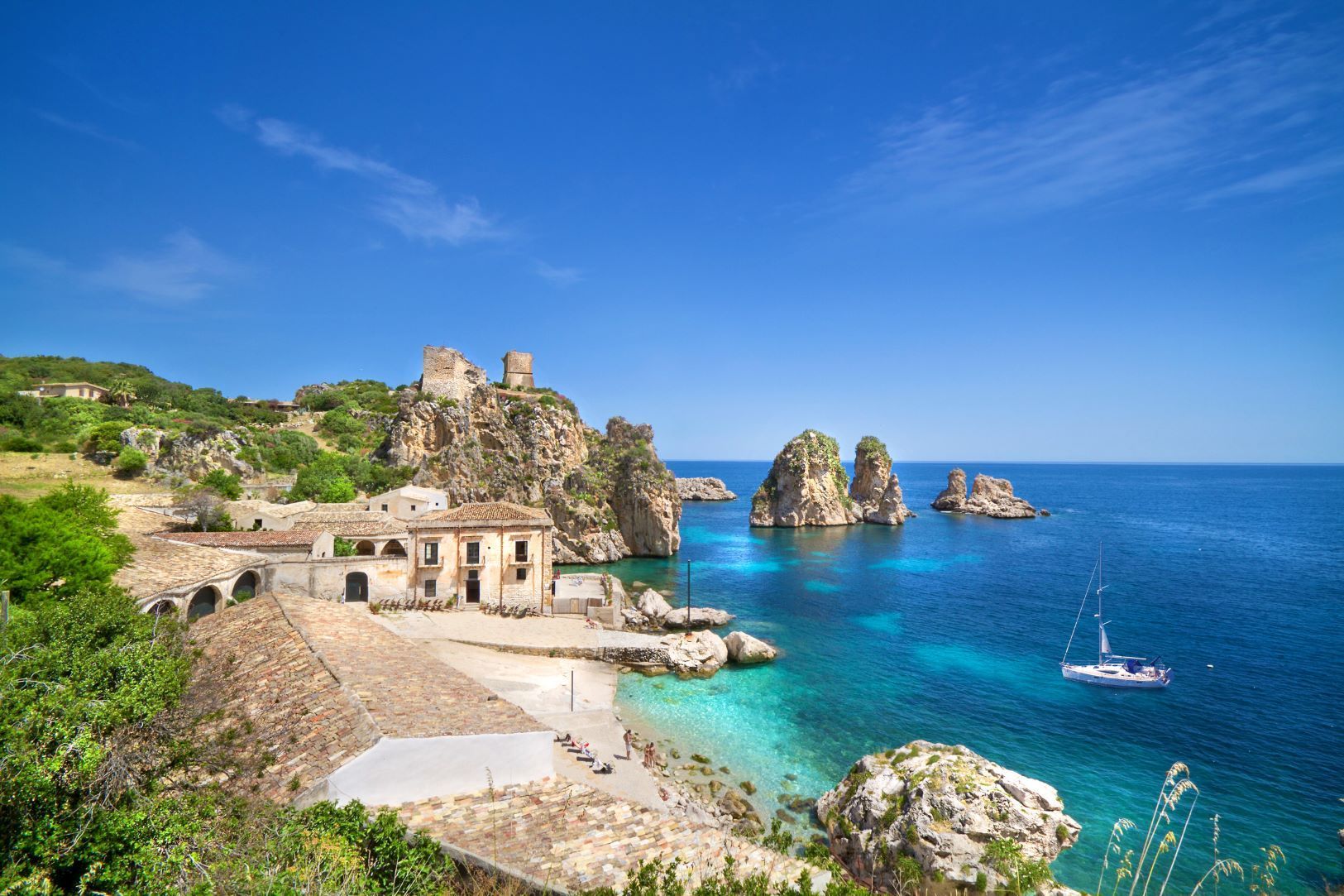 Un borgo costiero con le rovine di un castello su una scogliera, che si affaccia su una baia turchese con una barca a vela e grandi formazioni rocciose.