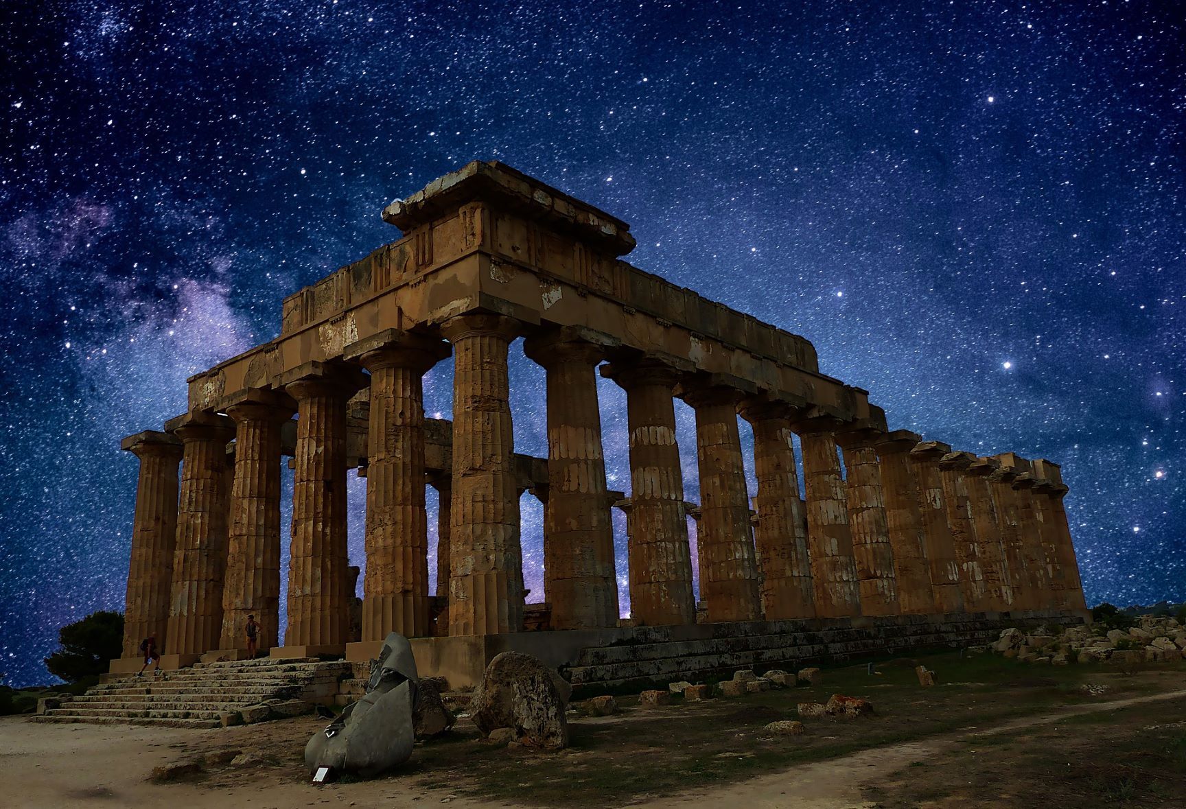 Le rovine di un antico tempio con grandi colonne di pietra si ergono illuminate contro un vivace cielo notturno pieno di stelle.