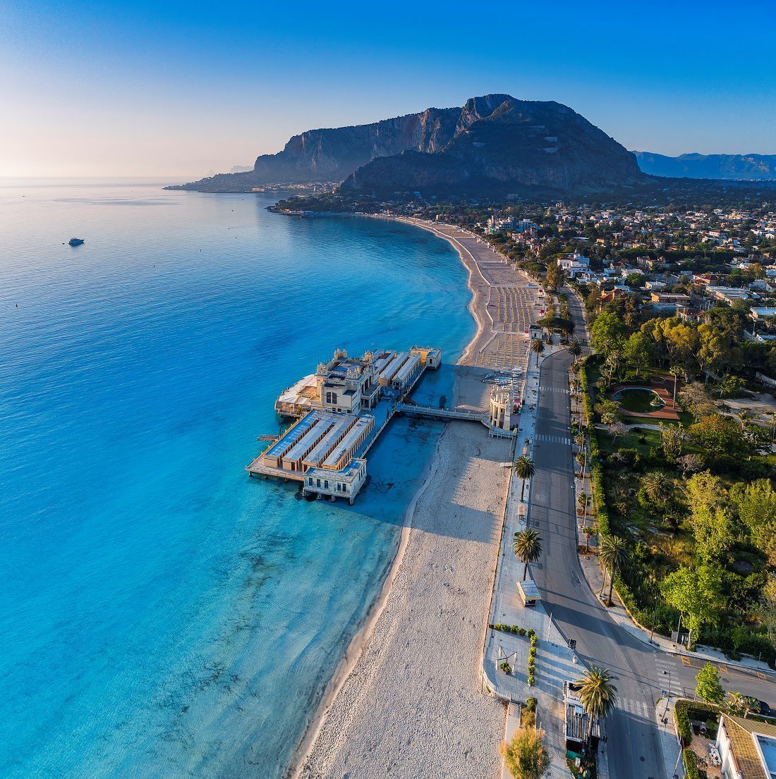 Vista aerea di una lunga spiaggia sabbiosa con un molo ornato che si protende nel mare turchese, accanto a una città ai piedi di una montagna.