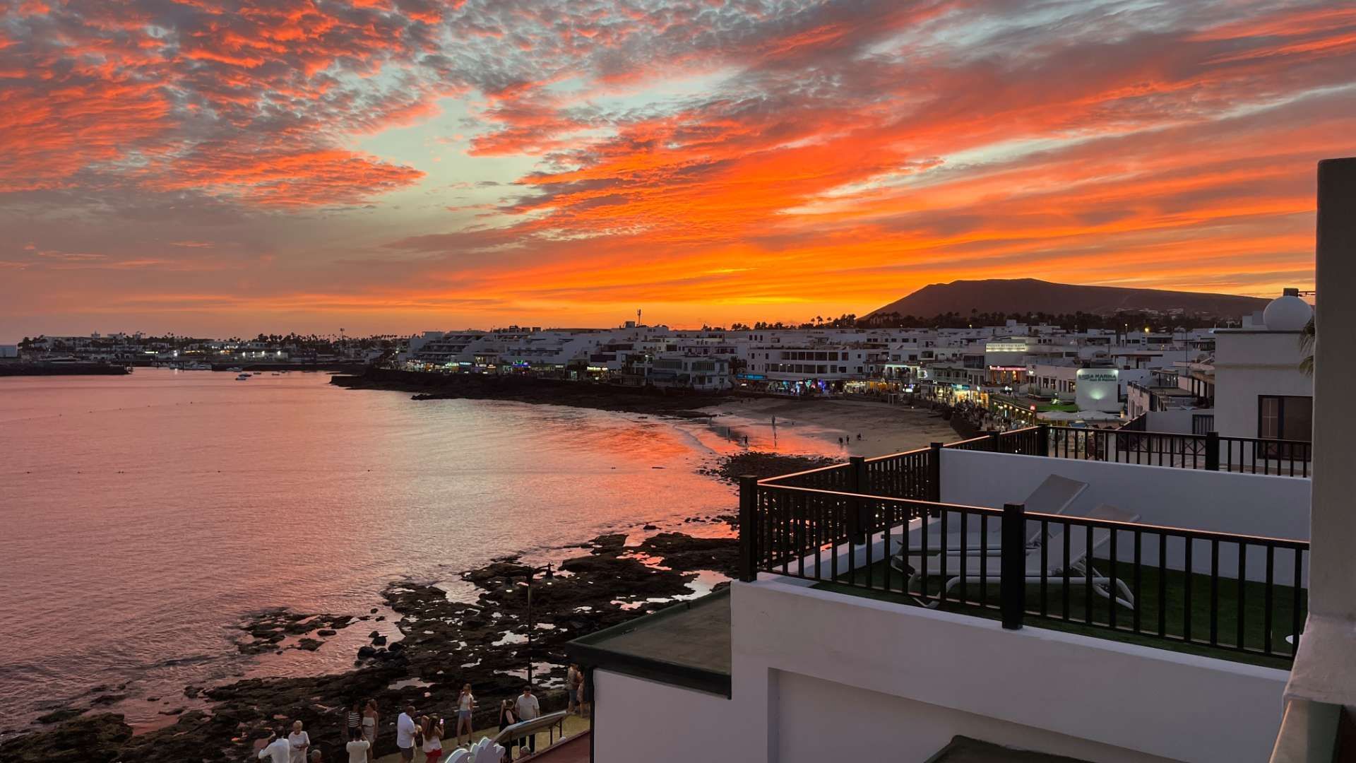 Città costiera con edifici bianchi, vista da un balcone sotto un cielo spettacolare arancione e rosso al tramonto.