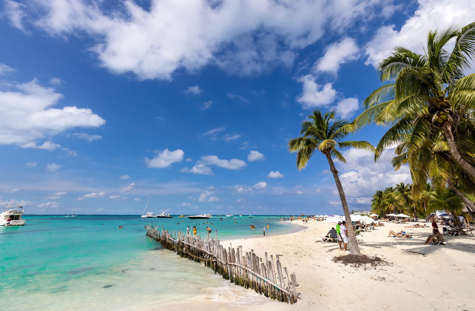 Una spiaggia tropicale soleggiata con palme, sabbia bianca e acqua turchese. Le persone si stanno rilassando e nuotando, con barche ancorate al largo.