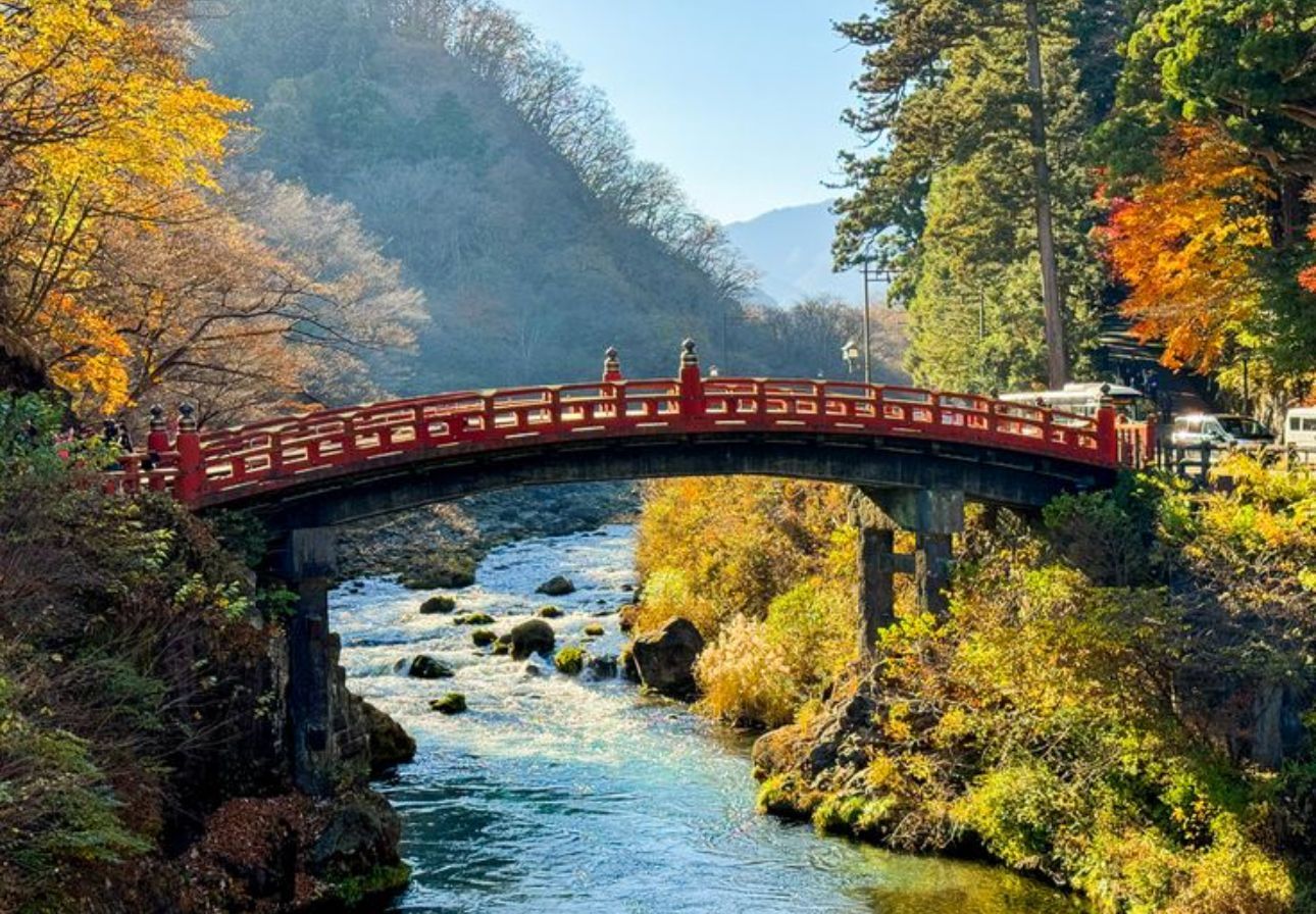 Un tradizionale ponte ad arco rosso attraversa un fiume limpido e roccioso in una valle con alberi autunnali colorati.