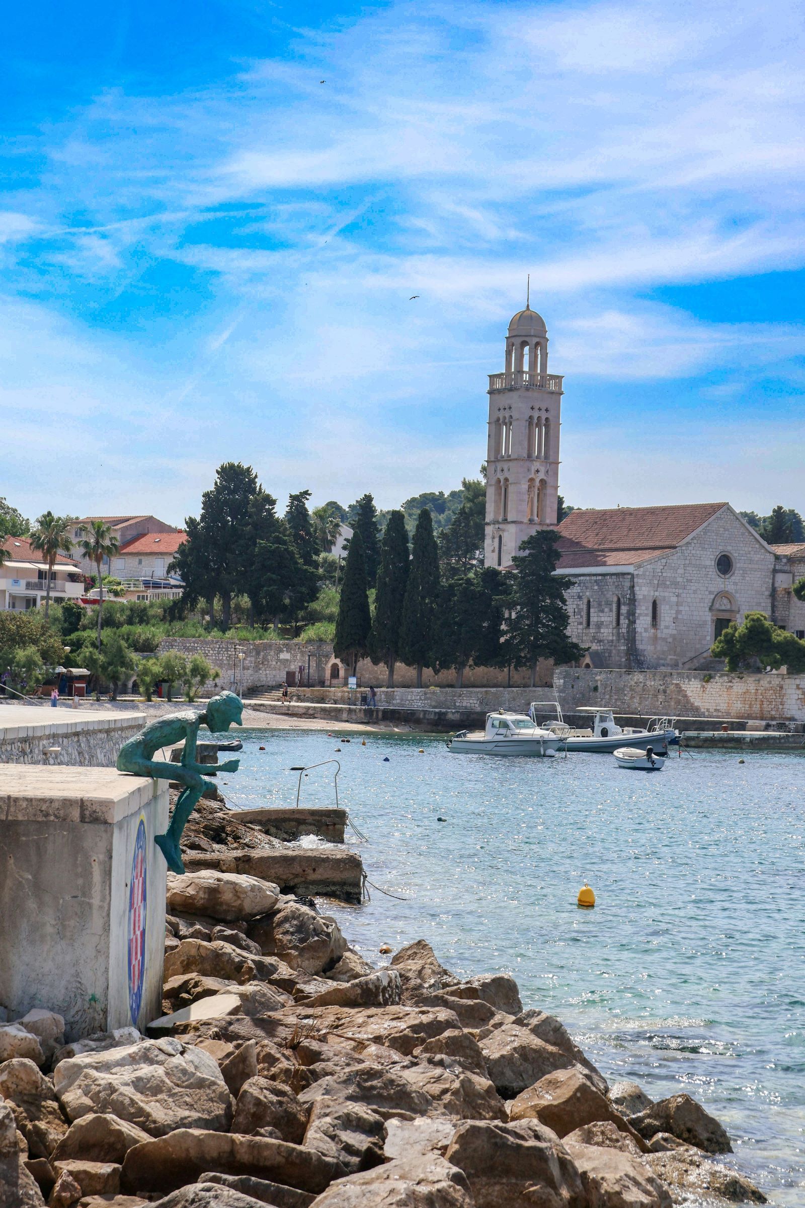 Una statua di bronzo si trova su un molo di cemento che si affaccia su una baia tranquilla con barche e una chiesa in pietra con campanile.