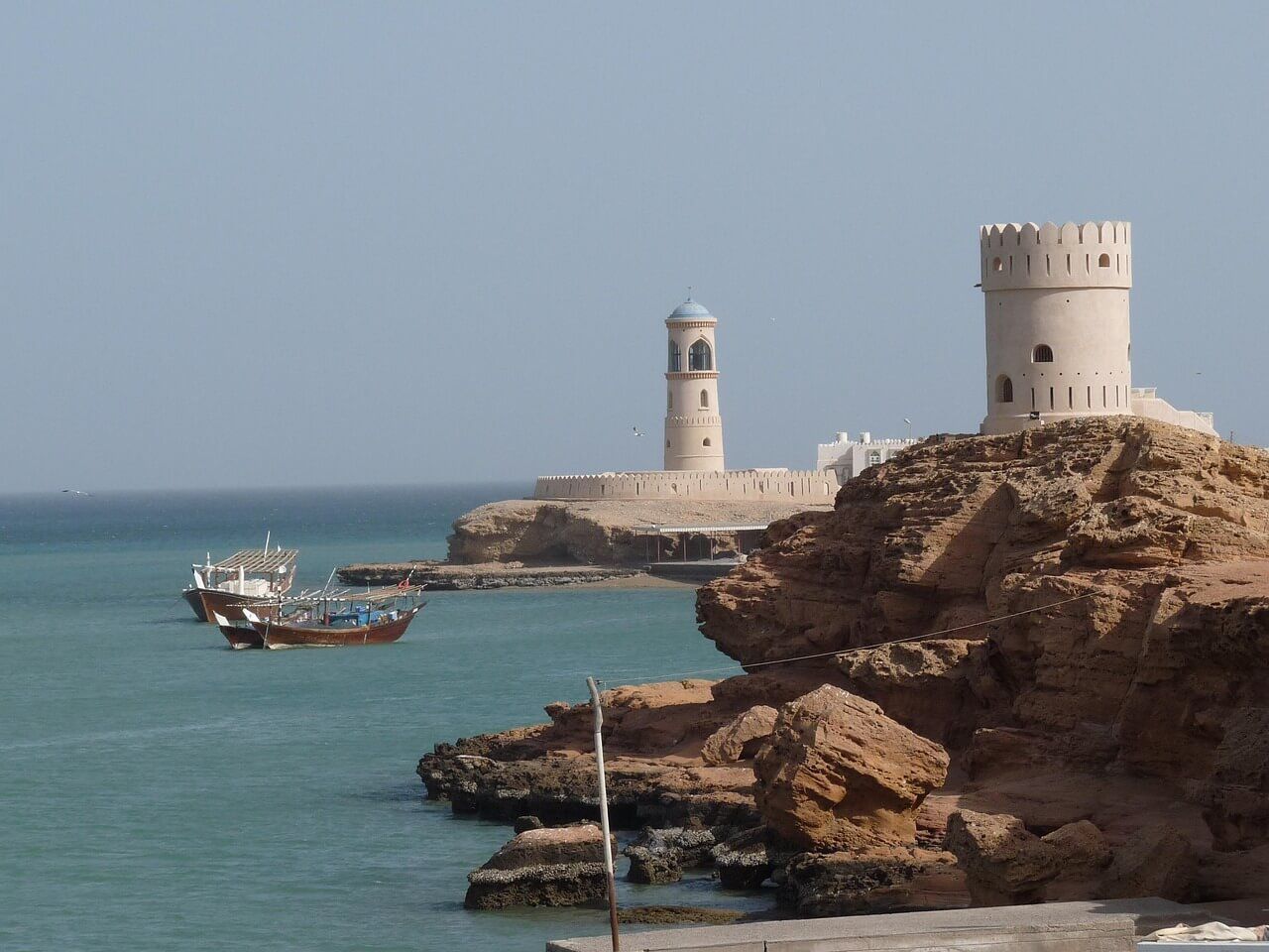 A lighthouse and a watchtower on a rocky coastline overlook a calm bay with two traditional boats.