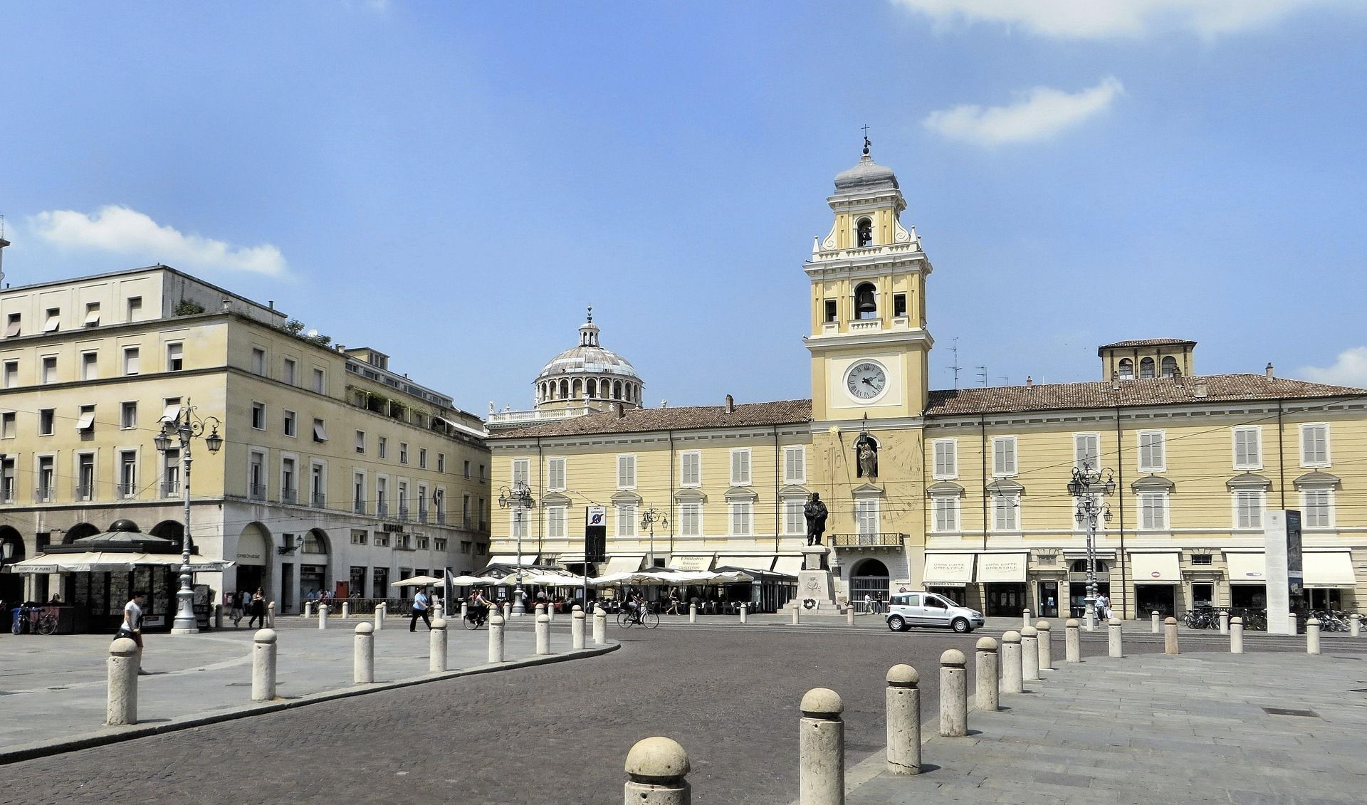 Un'ampia piazza europea acciottolata, affiancata da storici edifici gialli, con un'imponente torre dell'orologio e una cupola.