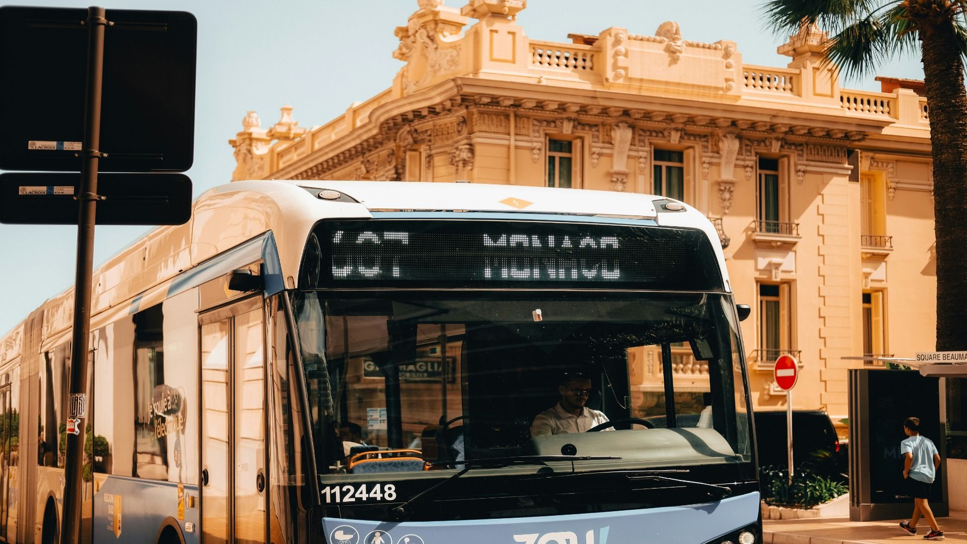 Vista frontale di un autobus blu e bianco su una strada cittadina, con un autista al volante e un edificio giallo sullo sfondo.