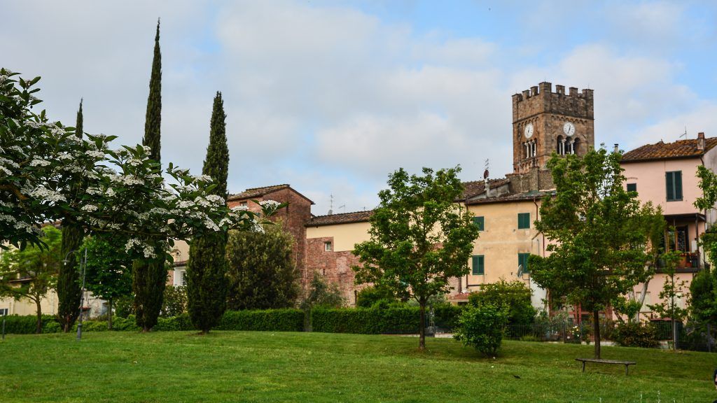 Un parco erboso con cespugli fioriti e cipressi di fronte a vecchi edifici, inclusa una prominente torre dell'orologio in pietra.