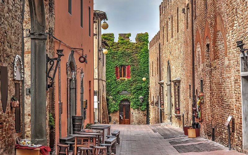 Uno stretto vicolo in mattoni con tavoli da caffè all'aperto conduce a un edificio storico coperto di edera verde.