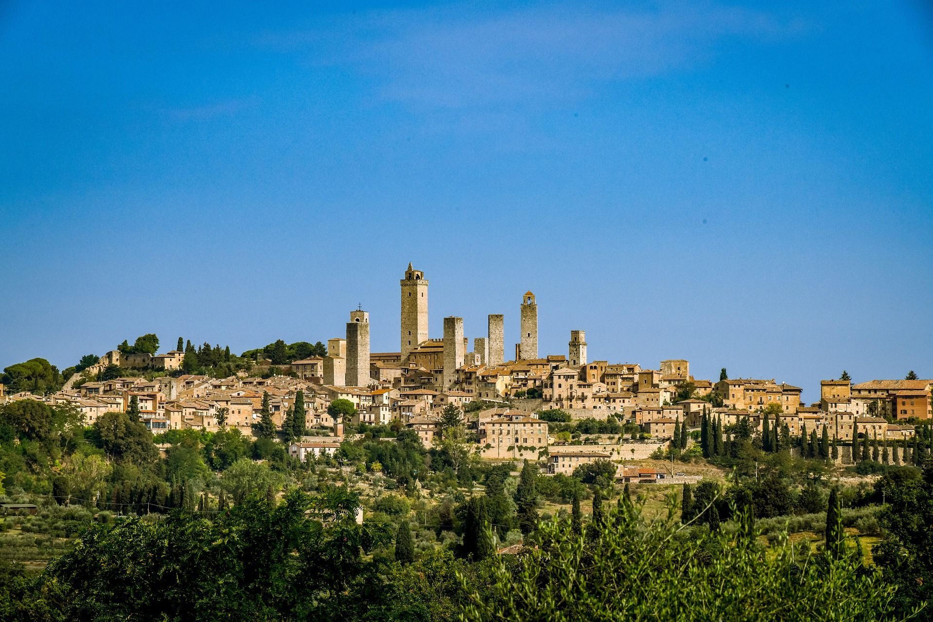 Una città storica con molteplici torri in pietra sorge su una collina verde lussureggiante, circondata da alberi sotto un cielo azzurro limpido.
