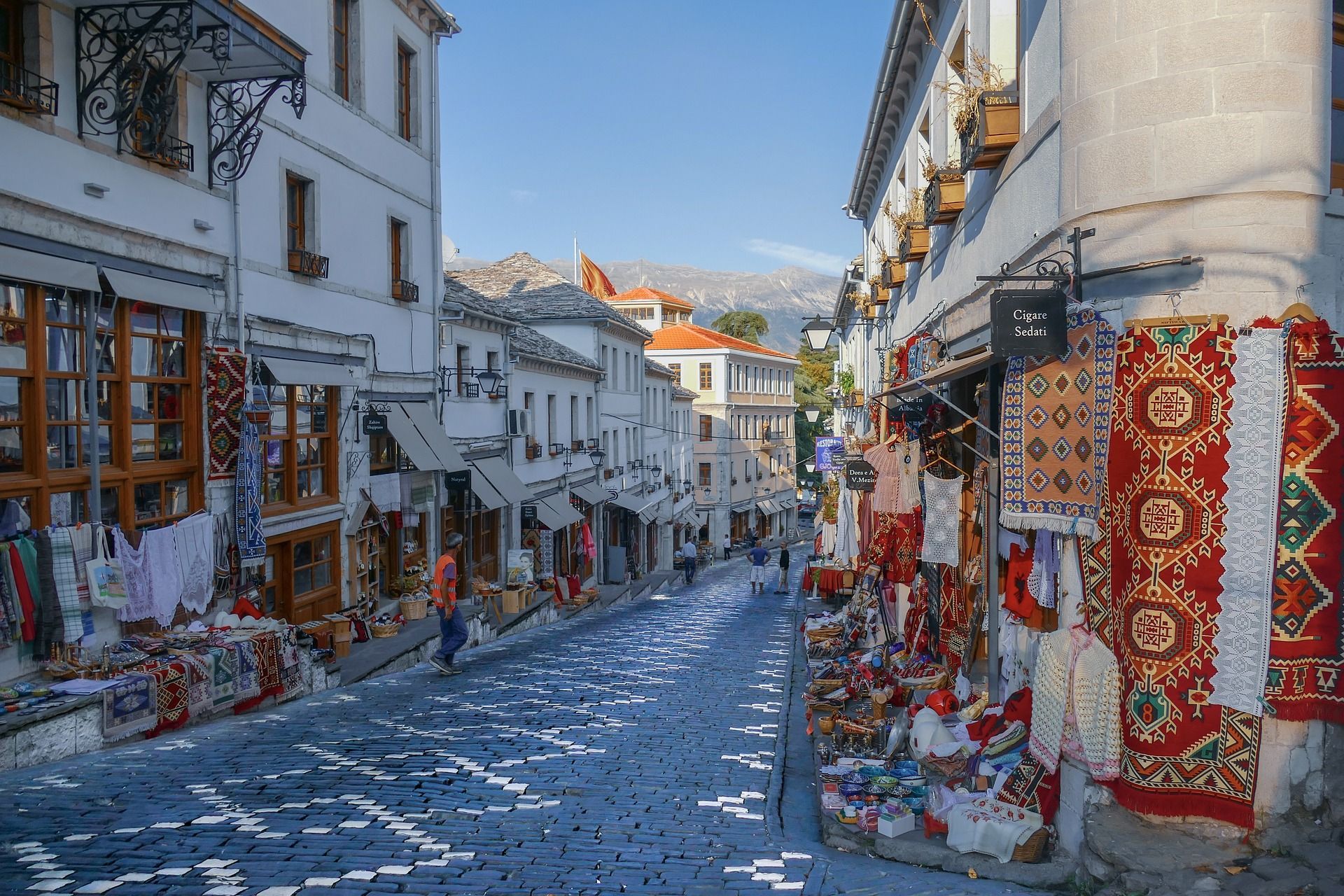 Una vista de una calle adoquinada con patrones, bordeada por tiendas que exhiben alfombras y textiles coloridos, con montañas al fondo.
