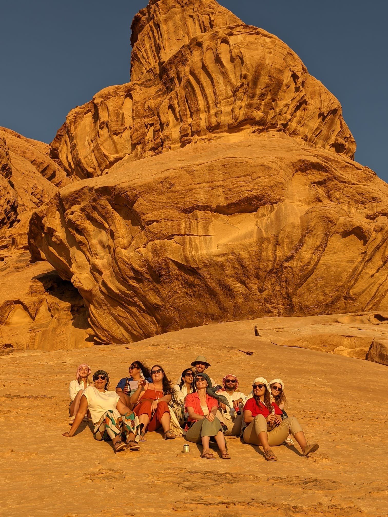 A WeRoad group trip sits together on a vast rock formation in a desert, with a towering cliff behind them.
