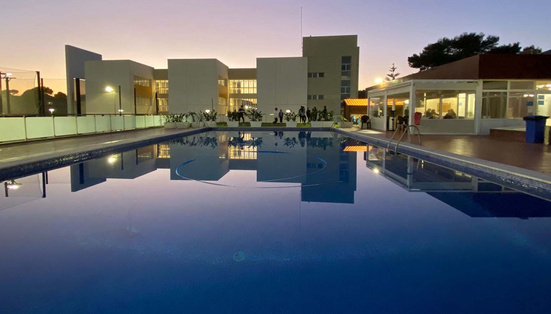 Una piscina riflette un edificio moderno e le luci al calar della sera, con un gruppo WeRoad riunito sul bordo piscina.