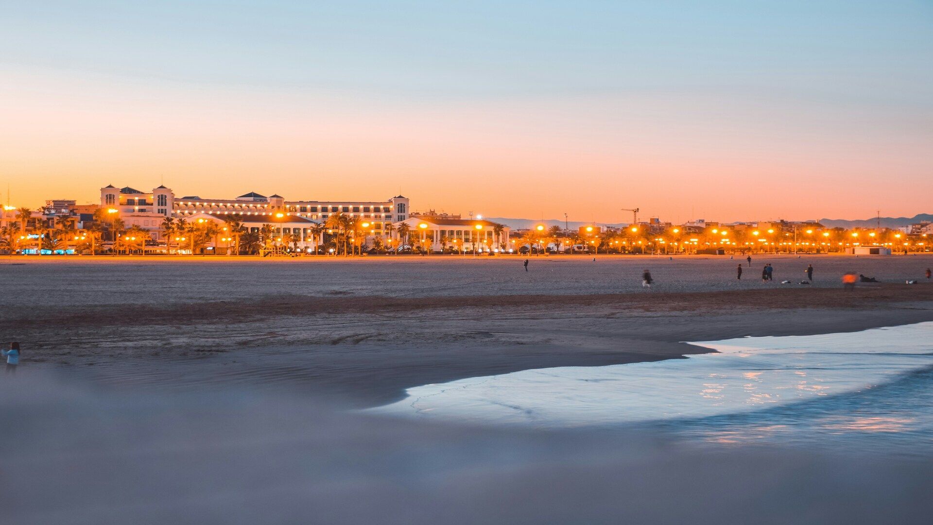 Ampia spiaggia sabbiosa al tramonto, con la battigia in primo piano e uno skyline cittadino illuminato sotto un cielo arancione e blu.