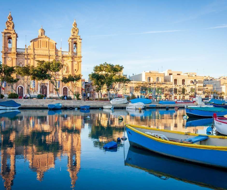 Una grande chiesa in pietra con due campanili si riflette nelle acque calme di un porto pieno di piccole barche colorate.