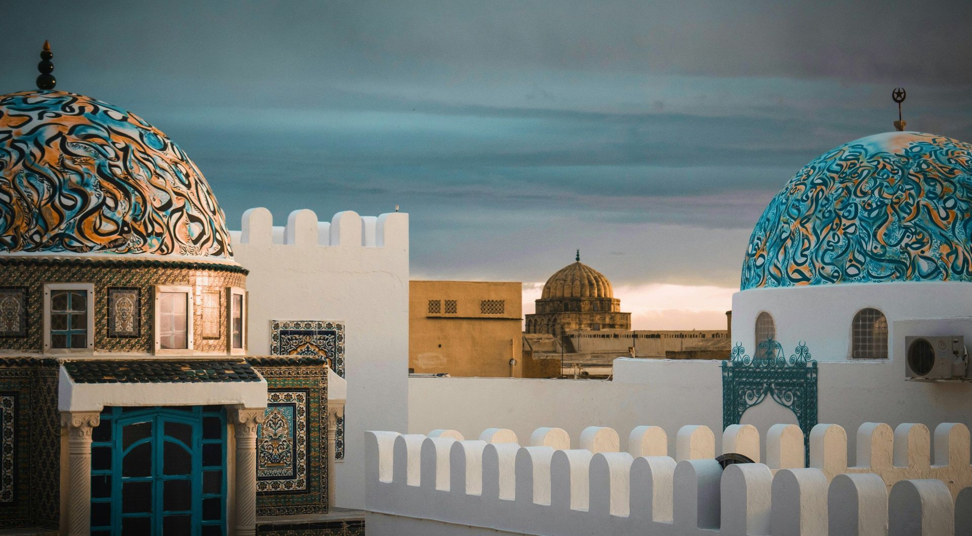 Due cupole riccamente decorate blu e arancioni svettano su edifici imbiancati a calce con mura merlate sotto un cielo nuvoloso.