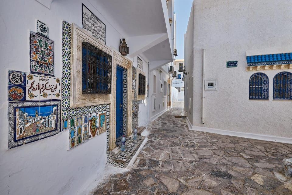A narrow cobblestone alley between whitewashed buildings, one decorated with a blue door and colorful, ornate ceramic tiles.