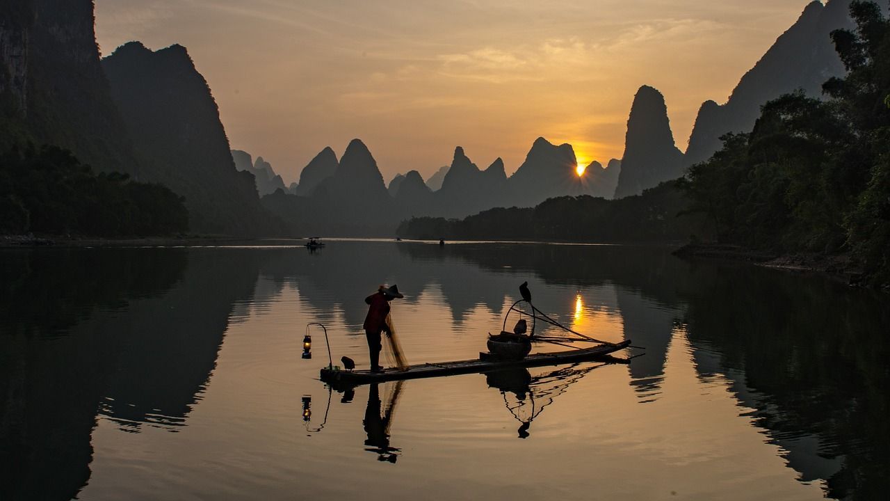Un pescador en una balsa de bambú lanza una red en un río tranquilo al amanecer, con cormoranes y montañas silueteadas.