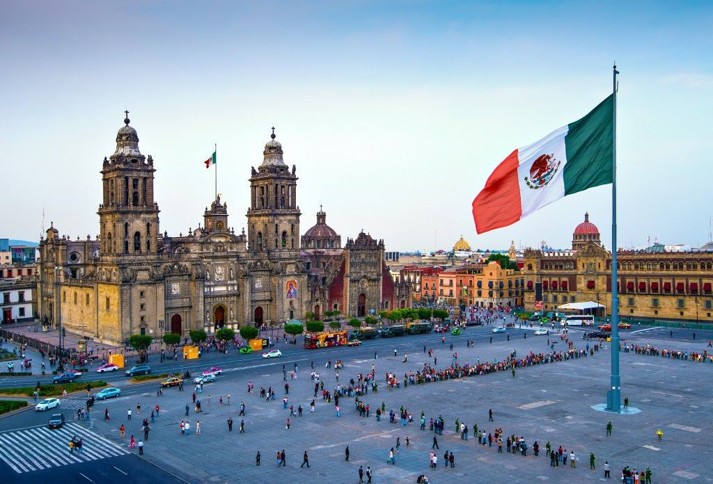 A large Mexican flag waves over a crowded city square in front of a historic cathedral building.
