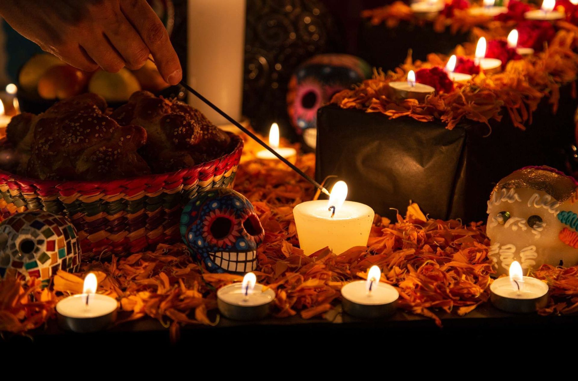 A hand lights an incense stick over a candle-lit altar with sugar skulls, pan de muerto, and marigold petals for Día de los Muertos.