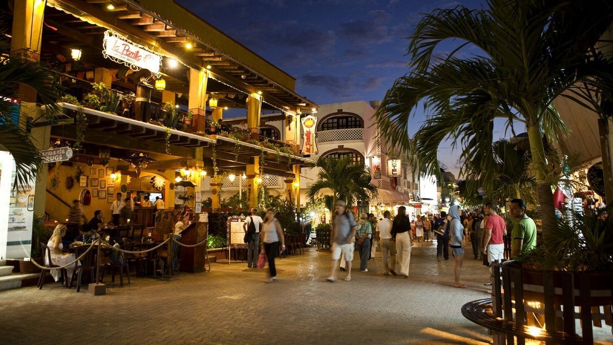 People walk along a busy cobblestone street at dusk, past illuminated multi-story restaurants with outdoor seating and palm trees.