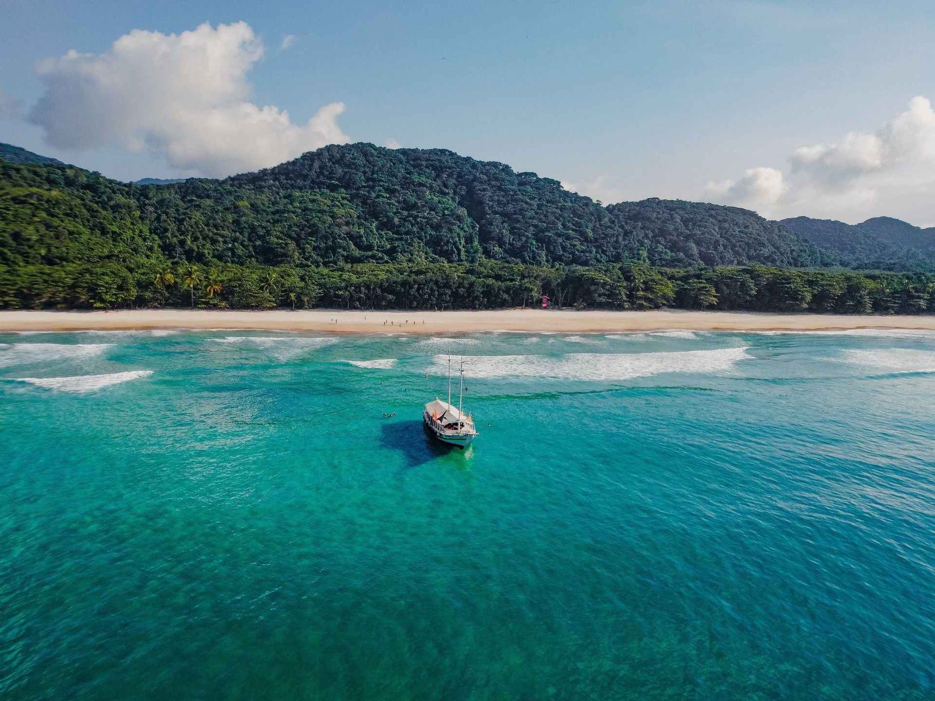 Una vista aérea de un barco blanco anclado en agua turquesa, con una playa de arena y una exuberante montaña verde al fondo.