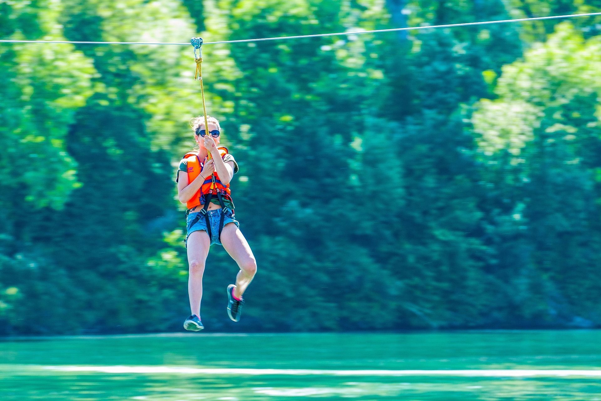 Una donna con giubbotto di salvataggio e occhiali da sole sfreccia in zip-line sopra uno specchio d'acqua, di fronte a una foresta verde.