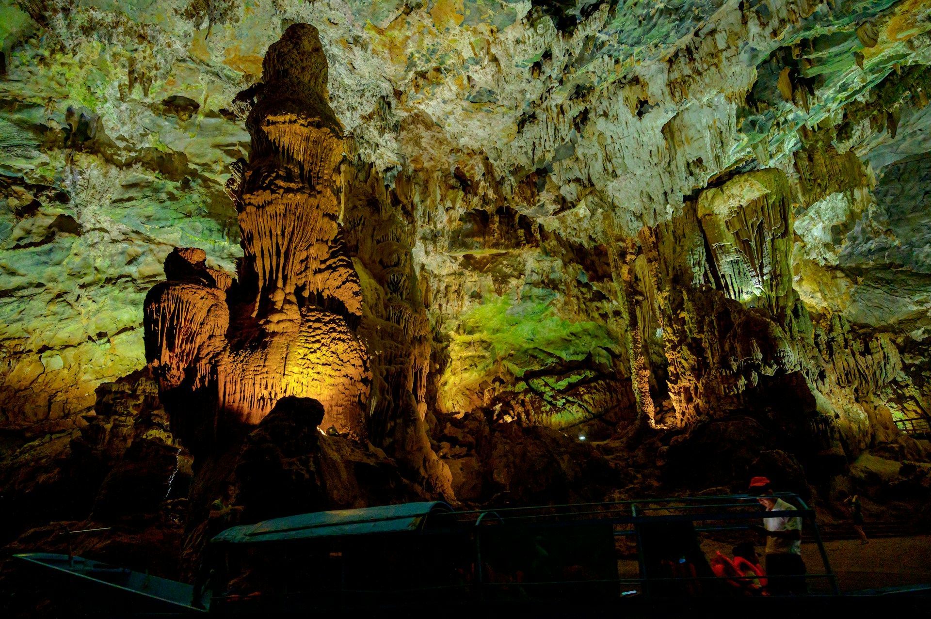 Una barca con un gruppo WeRoad naviga attraverso un'immensa caverna, con stalattiti e stalagmiti illuminate da luci verdi e gialle.