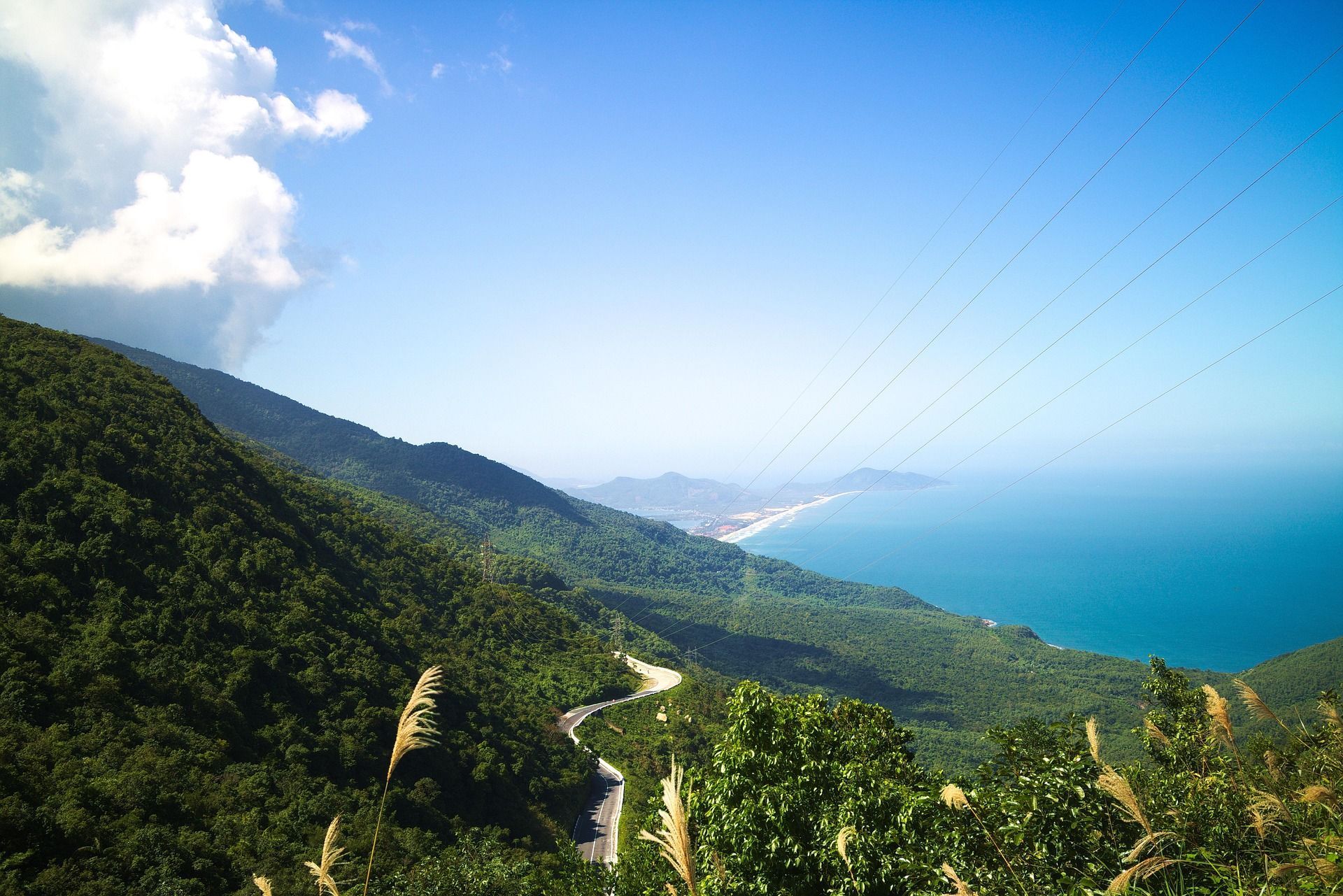 Vista panoramica di una strada tortuosa su una montagna verdeggiante che domina una costa sabbiosa e il mare blu.