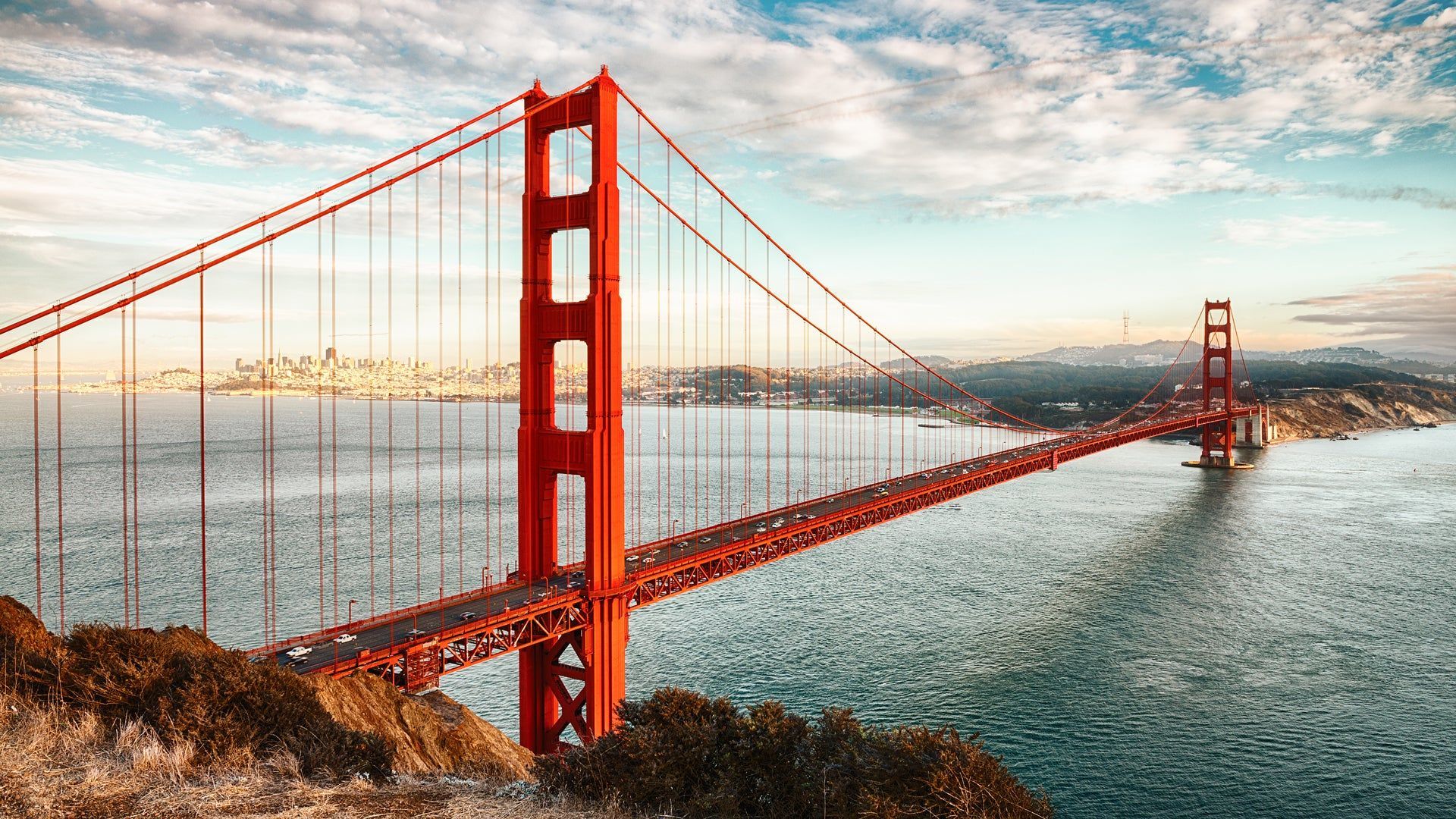 Una vista panoramica del Golden Gate Bridge che attraversa l'acqua, con lo skyline della città in lontananza sotto un cielo parzialmente nuvoloso.