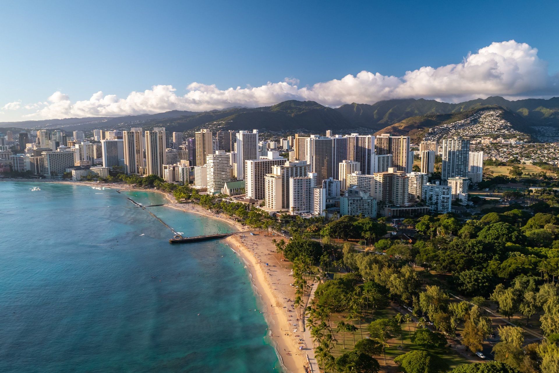 Una vista aerea dello skyline di una città lungo una spiaggia sabbiosa e un oceano turchese, con lussureggianti montagne verdi sullo sfondo.