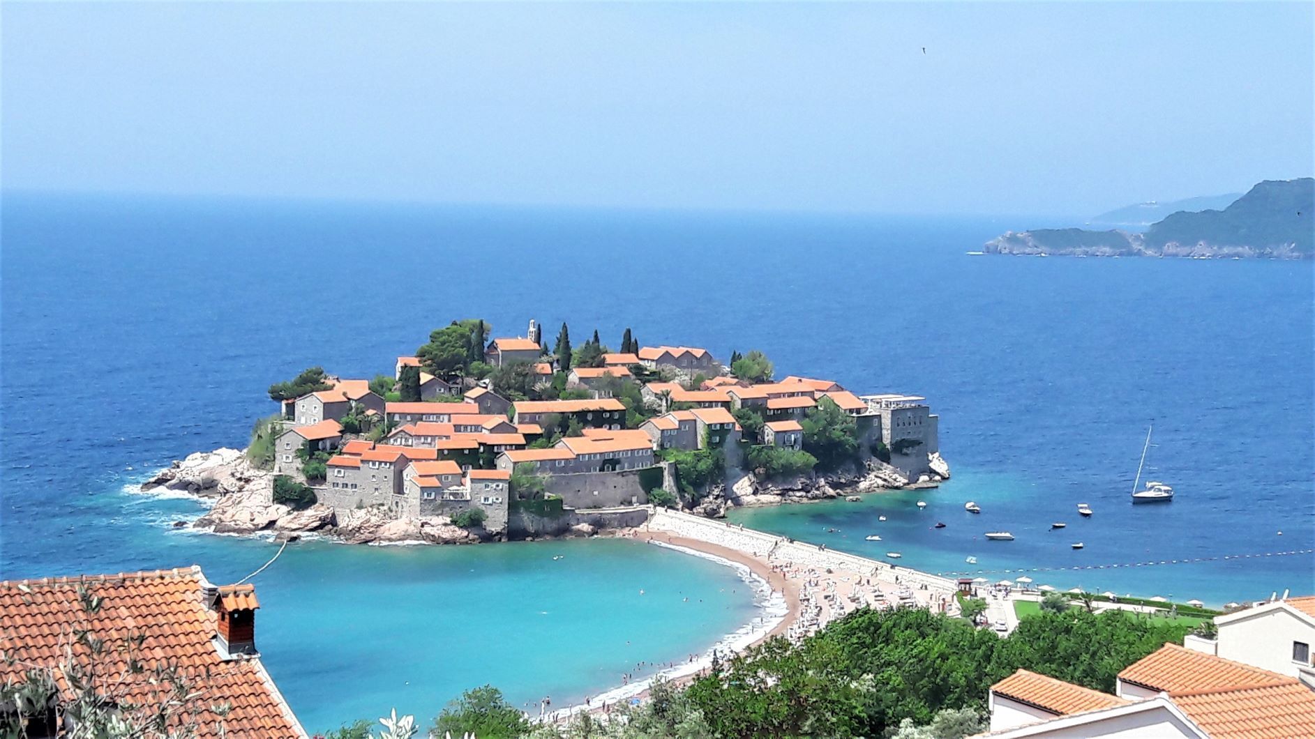Una piccola isola fortificata con edifici in pietra dai tetti rossi, collegata da un istmo a una spiaggia sabbiosa con acqua turchese.
