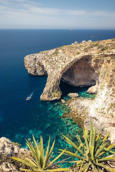 Vista dall'alto di una costa rocciosa con un grande arco naturale sull'acqua turchese, dove una piccola barca naviga.