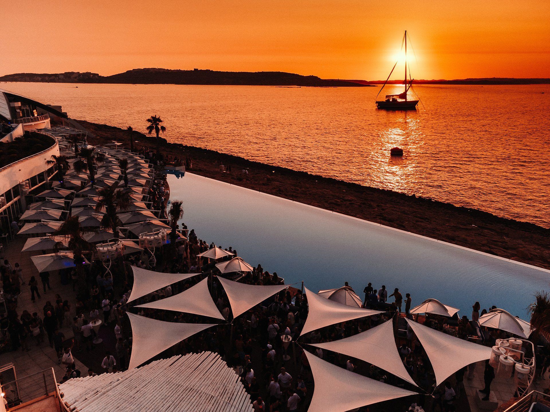 Una vista aerea di una festa affollata in una location sul mare con una piscina a sfioro, con una barca a vela sull'acqua durante un tramonto arancione.